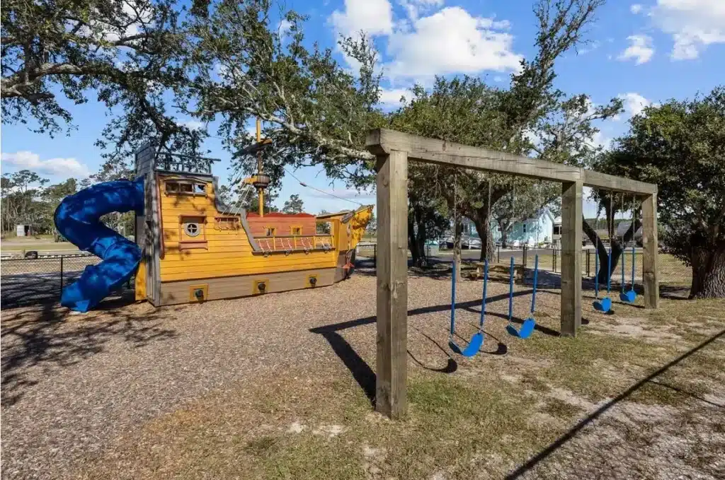 Outdoor playground with a pirate ship play structure, blue spiral slide, and wooden swing set on gravel under oak trees.