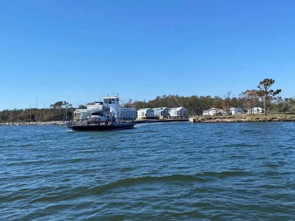 A vehicle ferry carrying a pickup truck crosses open blue water past waterfront stilt cottages and pine trees.