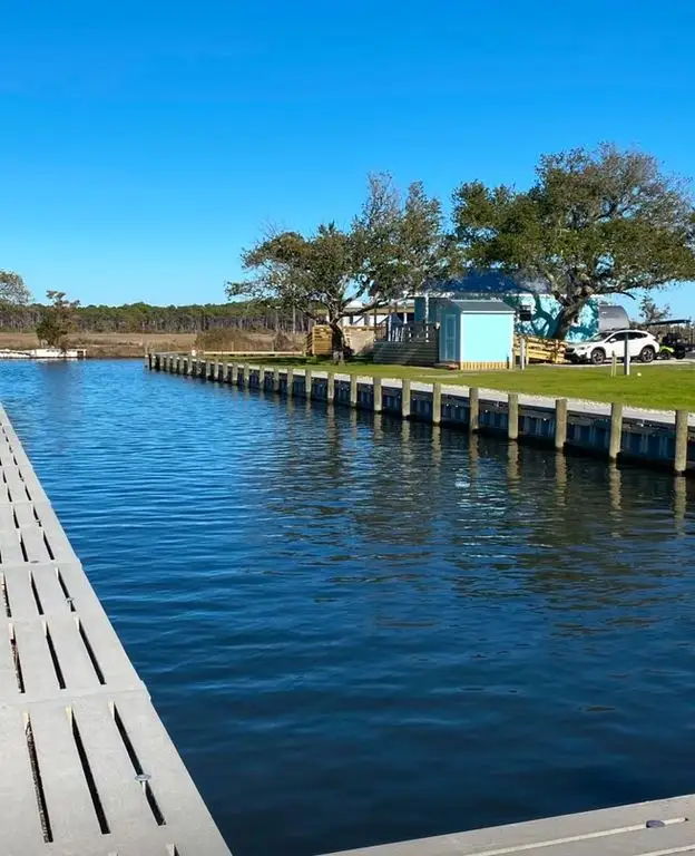 Calm canal lined with a wooden bulkhead, viewed from a floating dock, with a light blue shed, grassy lawn and parked