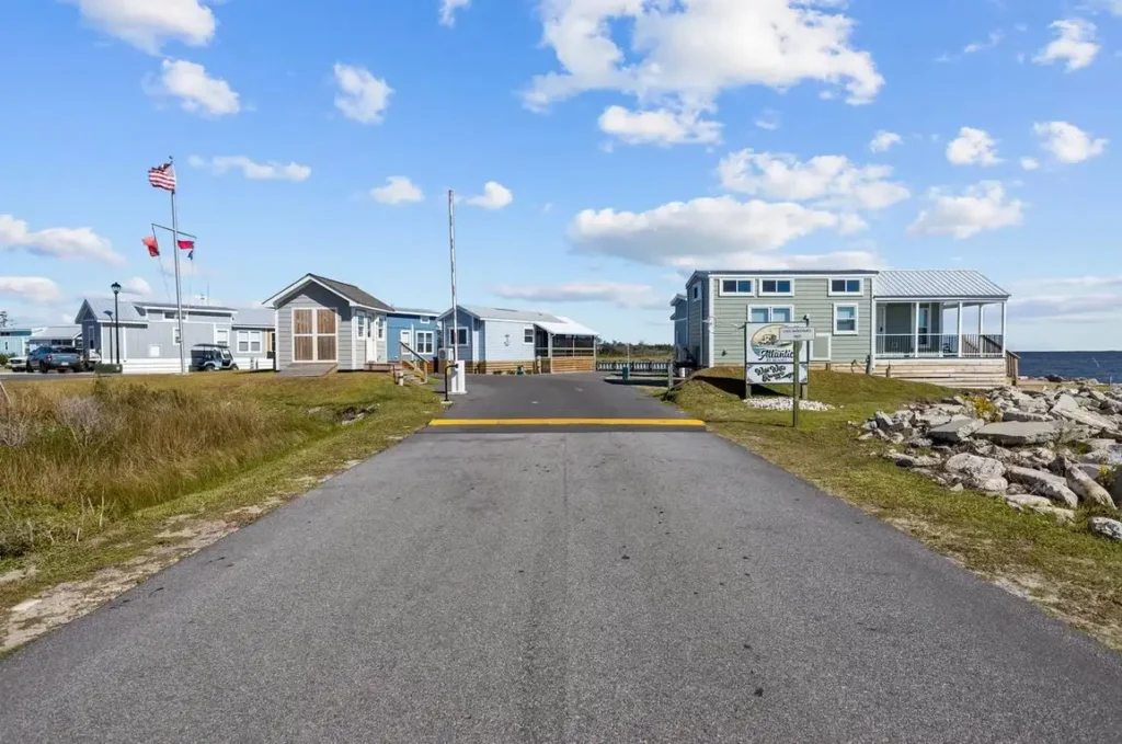 Paved entrance road leading into a waterfront RV park lined with cottages, an American flag, and a sign near rocky