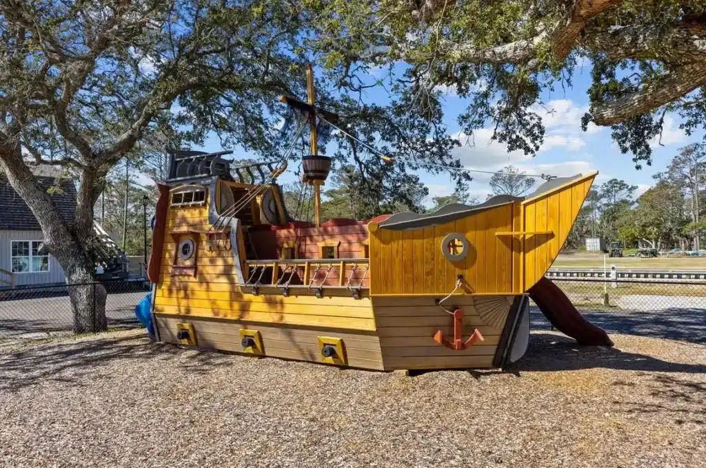 Wooden pirate ship playground structure with slide and crow's nest mast sits on gravel beneath large oak trees.