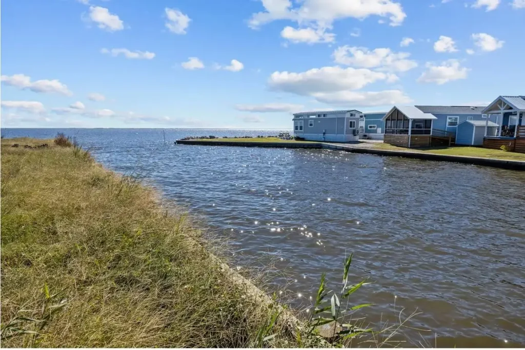 Waterfront canal beside grassy bank with a row of light blue park model cabins and screened porches under blue sky.