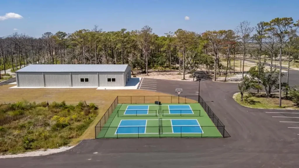 Aerial view of fenced green pickleball courts with basketball hoop beside a large gray metal building and paved parking