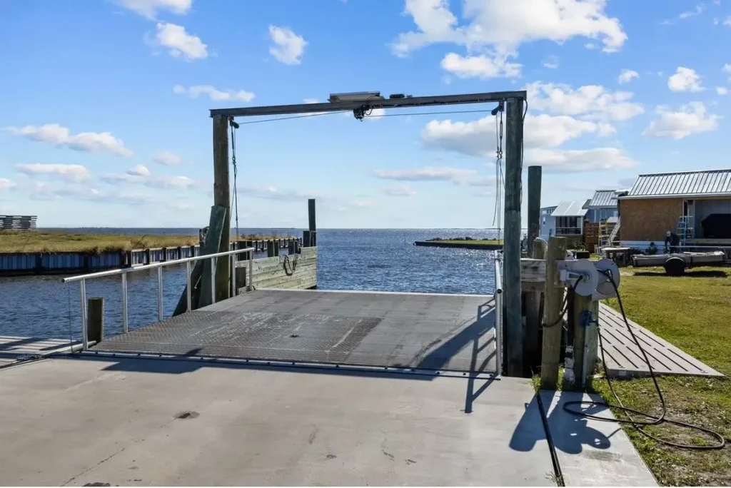 Concrete boat ramp and dock with metal boat lift frame, pilings, and hose reel beside open bay water.