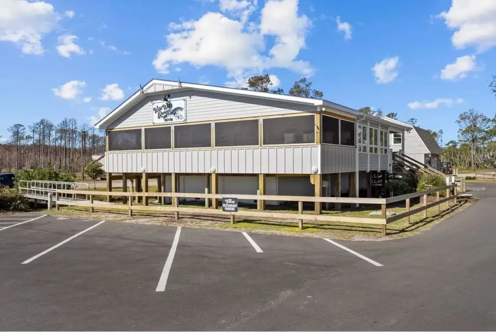 Elevated gray restaurant building on pilings with screened porch, wooden fence, and empty striped asphalt parking lot