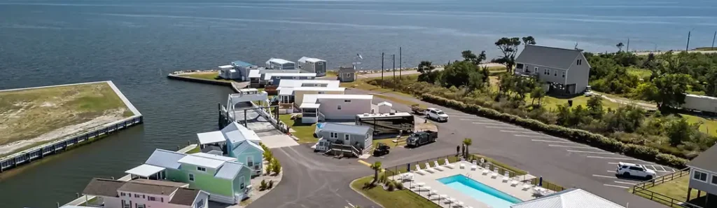 Aerial view of a waterfront RV resort with pastel tiny-home cabins, boat docks, paved parking lots, and a swimming pool