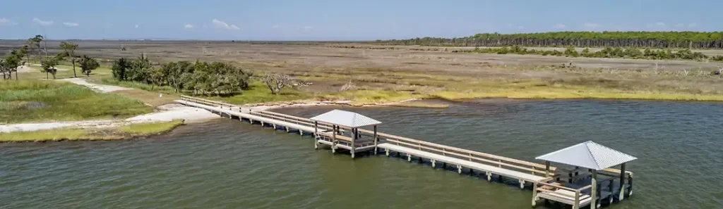 Aerial view of a long wooden fishing pier with two covered gazebos extending from a marsh shoreline into calm water.