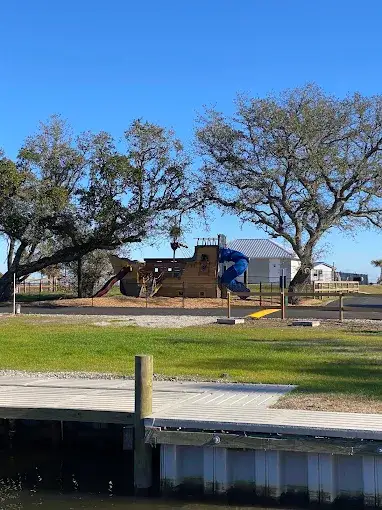 Wooden playground structure with blue slide under large oak trees, seen across a grassy lawn from a waterfront dock.