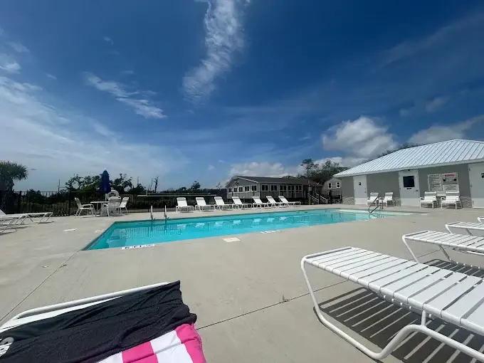 Outdoor swimming pool with white lounge chairs on concrete deck beside a metal-roofed pool house and clubhouse building.