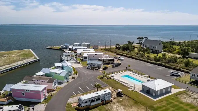 Aerial photo of a waterfront RV resort with pastel tiny-home cabins, swimming pool, bathhouse, parked travel trailers